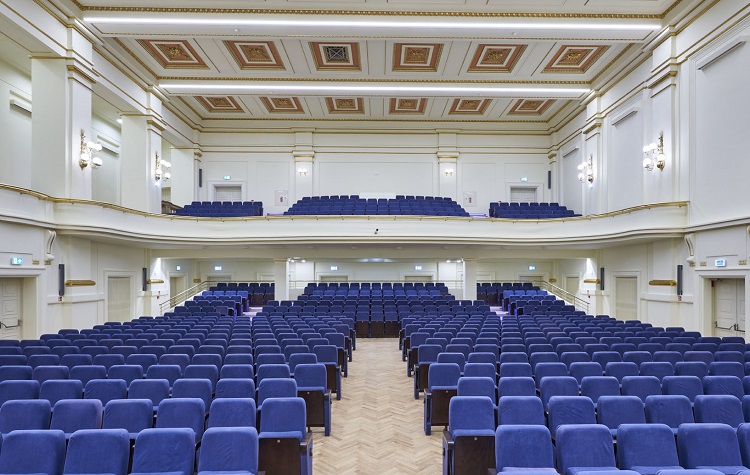 Duża, pusta sala koncertowa z rzędem niebieskich foteli. Widok na część widowni. Sala posiada dwa poziomy miejsc siedzących oraz elegancki wystrój w stylu klasycznym z ozdobnymi panelami na suficie. fot. P. Ulatowski